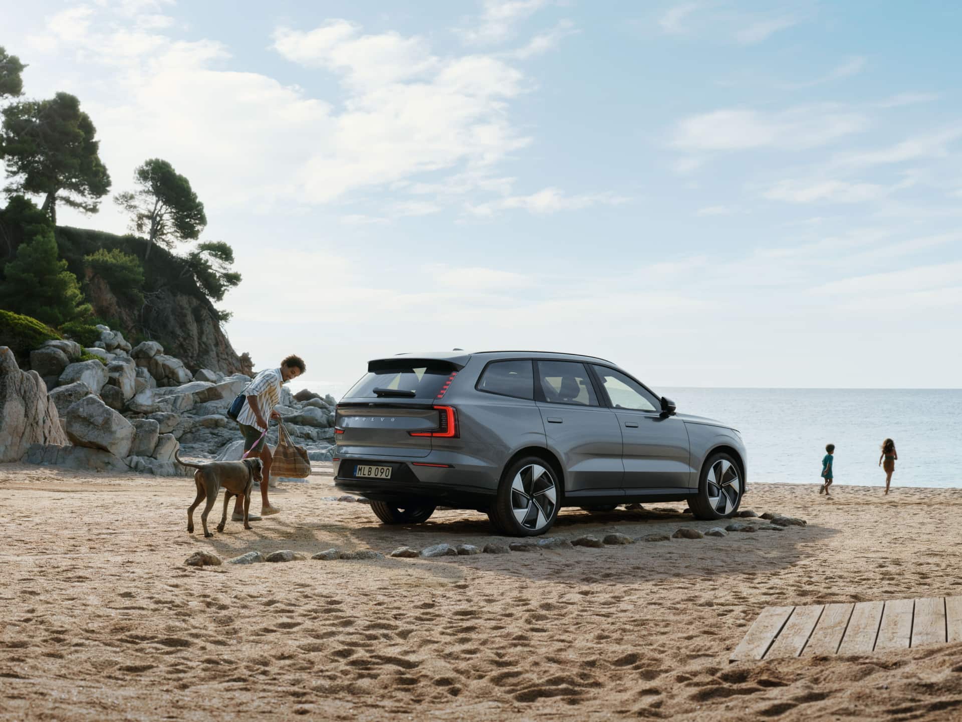Ein Bild, das einen Mann und einen Hund zeigt, die neben einem am Strand geparkten Volvo EX90 spazieren gehen.