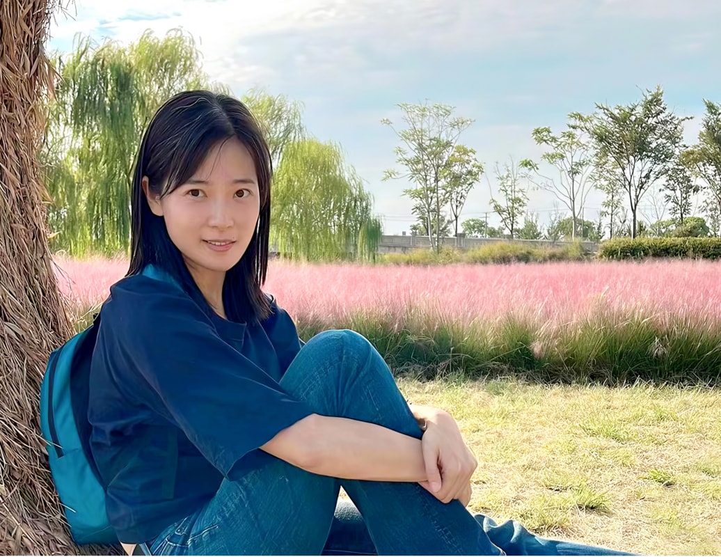 A young Asian woman sitting leaning against a tree in front of a field of pink muhly grass. 