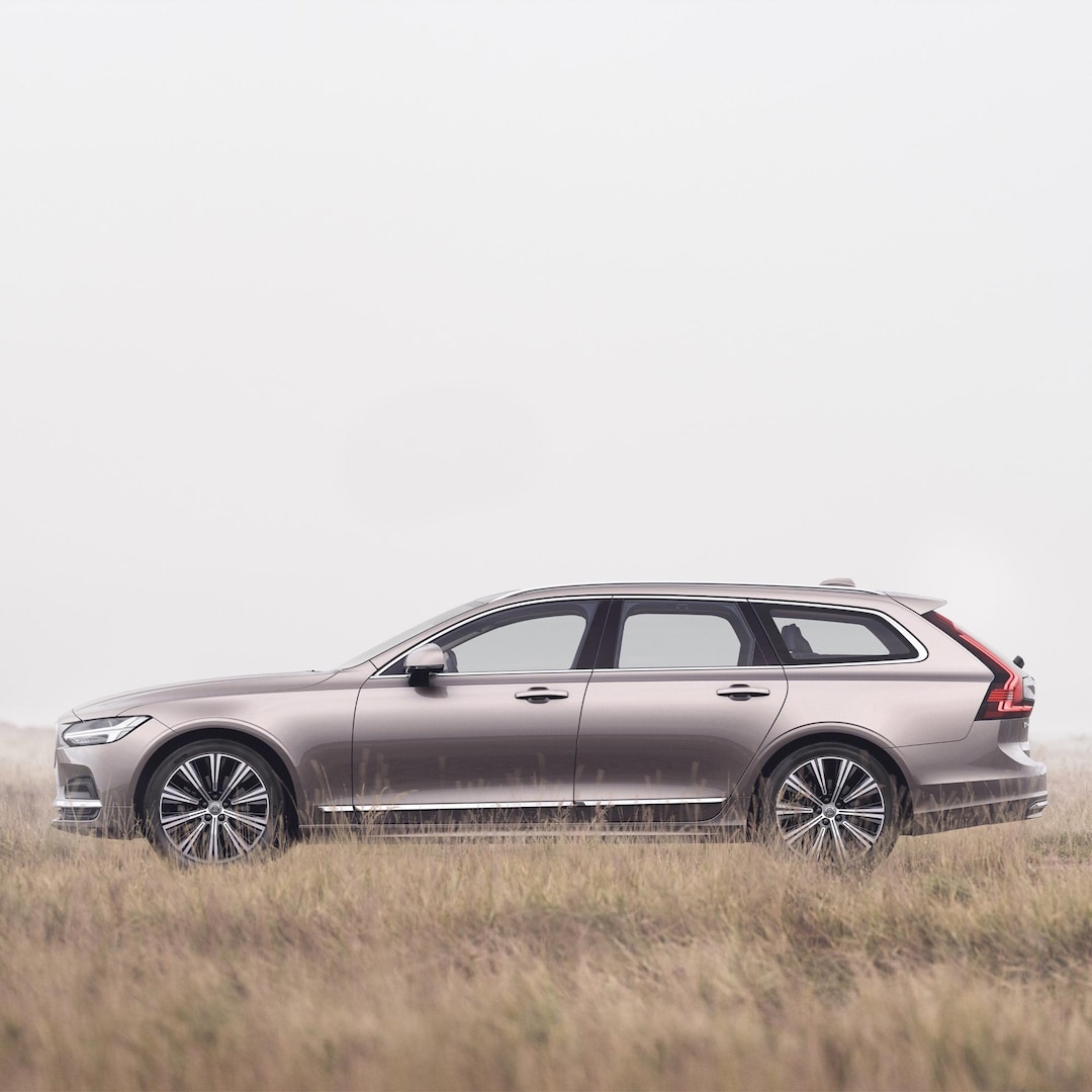 Weite Aufnahme eines Volvo V90-Kombis in Bright Dusk, der in einem nebligen Feld aus weichem, goldenem Gras geparkt steht.