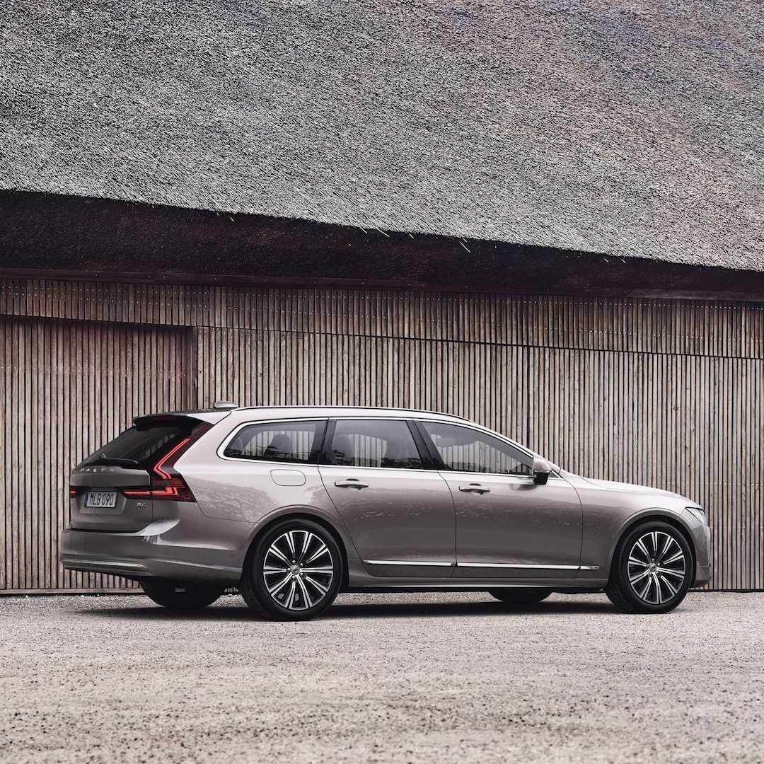 Ein Volvo V90 Kombi in Bright Dusk, geparkt in der Nähe der Holzaussenwände eines modernen Reetdachhauses.