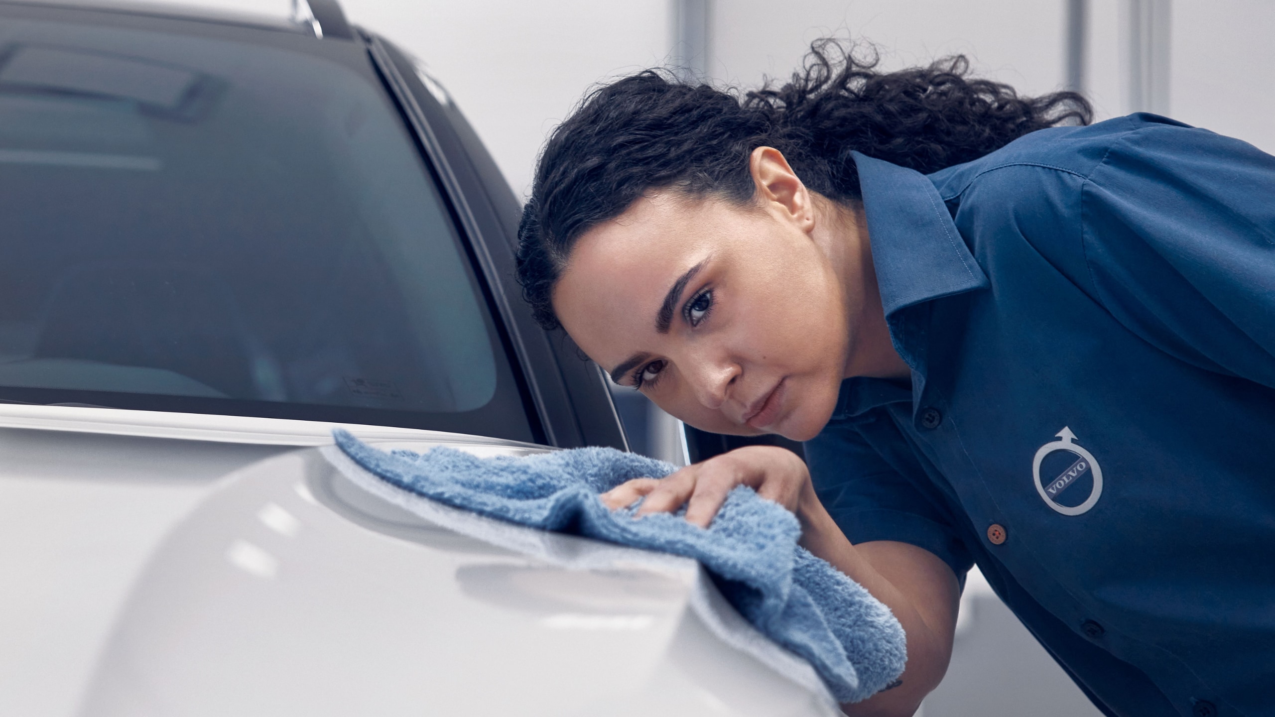 Eine Frau im blauen Volvo Poloshirt geht mit einem blauen Tuch über den weißen lack eines Volvo Fahrzeuges.